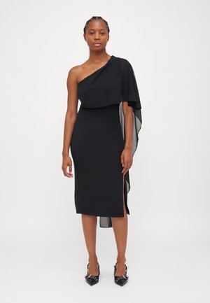 Robe noire avec un design asymétrique à une épaule, superposition en chiffon superposée, silhouette ajustée et fente sur le côté. Associée à des chaussures à talons noires.