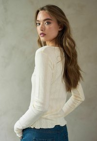 Krems krāsas garu piedurkņu tops ar ribotu tekstūru un viļņotu apakšmalu, apvienots ar zilajiem augstajiem džinsiem. Audumam ir maigas vertikālas svītras.