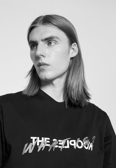 Schwarzes Baumwoll-T-Shirt mit kräftigem, stilisiertem weißen und grauen Text, der auf der Vorderseite "DIE OBJEKTE? WAS IST?" lautet. Einfacher Rundhalsausschnitt.