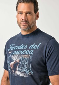 Man met kort donker haar en baard, gekleed in een marineblauw shirt met een afbeelding van een beer en een bos en de tekst "Fuentes del Narcea El Parque Natural".
