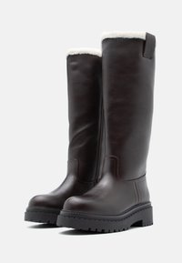 Bocanci din piele maro, până la genunchi, cu o căptușeală pufoasă albă, suprafață texturată, talpă groasă neagră și detalii de cusătură subtile pe cusături.