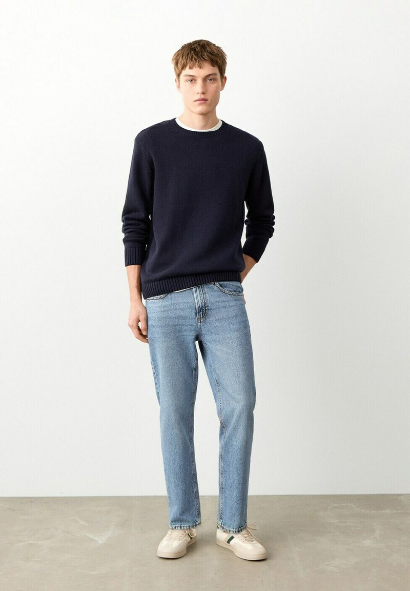 Jeune homme debout, une main dans la poche, portant un pull marine, un jean bleu clair et des baskets beige, devant un mur blanc uni.