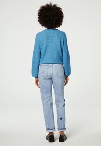 Niebieski sweter robiony na drutach w splot kablowy, zestawiony z jasnoniebieskimi dżinsami o prostym kroju z haftem gwiazdek. Całość dopełniają czarne buty z ćwiekami.