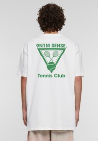 Balts īsām piedurknēm T-krekls ar aizmugurē izsistītu za�ļu grafiku, kurā attēloti tenisa racketi un bumba, ar uzrakstu "Tennis Club".