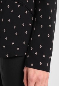 Svart tygblus med en texturerad yta och ett diamantsmönster i ljusrosa. Närbild av ärmen som visar mönsterdetaljen.