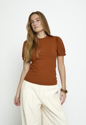 Moteris su ilgais šviesiai rudais plaukais, dėvinti rūdžių spalvos trumpą pūstą rankovę turinčią palaidinę ir plačias baltas kelnes, pozuojanti priešais vientisą baltą foną.