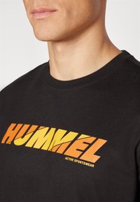 Juodas medvilnės marškinėliai su dideliu oranžiniu ir geltonu "HUMMEL" logotipu, su apvalia iškirpte ir trumpomis rankovėmis, skirti aktyviam dėvėjimui.