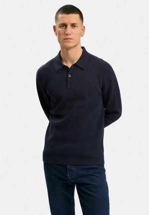 Mörkblå stickad polo-tröja med platt krage, tvåknapps öppning och långa ärmar; bärs med mörka jeans.