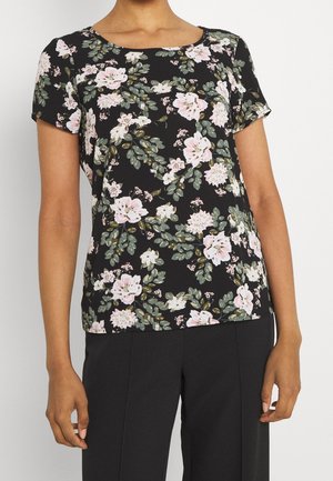 Femme portant un haut noir à manches courtes avec un motif floral rose et blanc et un pantalon noir, debout devant un fond uni.