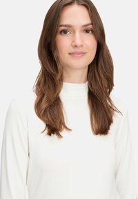 Biała bluzka z długim rękawem i wysokim kołnierzem, wykonana z miękkiego, gładkiego materiału o dopasowanej sylwetce i minimalnych detalach szwów.