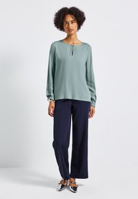 Blusa verde chiaro a maniche lunghe con scollatura a girocollo e dettaglio a chiave. Indossata con pantaloni larghi blu navy e scarpe piatte argento, su uno sfondo neutro.