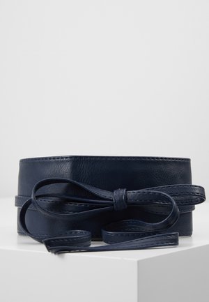 Large ceinture en cuir bleu marine avec un long nœud noué, présentée sur un piédestal blanc sur un fond uni.