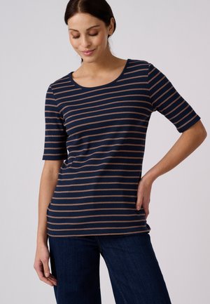 Mujer que lleva una camiseta azul marino de manga corta con finas rayas horizontales naranjas y vaqueros azul oscuro, de pie con una mano en la cadera.