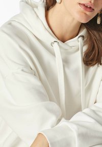 Witte capuchontrui gemaakt van zachte stof, met een relaxte pasvorm, zilveren oogjes en trekkoordjes. Close-up van de halslijn en mouwen.