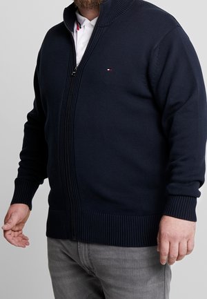 Mand iført navy lynlås-sweater over hvid skjorte med krave og grå bukser, stående med arme afslappet ned langs siderne.