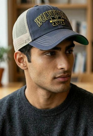 Junger Mann mit einer blauen und beigen Baseballkappe mit der Aufschrift "Wolverines Michigan" und einem dunklen Pullover, schaut leicht nach rechts drinnen, unscharfer Hintergrund.