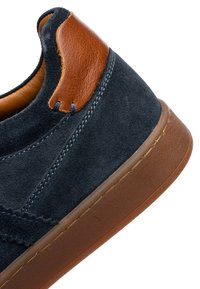 Tmavě modrá semišová teniska s hnědým koženým detailem na patě a hnědou gumovou podrážkou, zobrazená z blízkého bočního úhlu.
