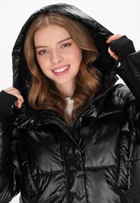 Fekete puffkabát fényes, ripstop bevonattal. Kapucnival, cipzárral és patentos záródással rendelkezik, rugalmas mandzsettákkal és steppelt mintázattal.