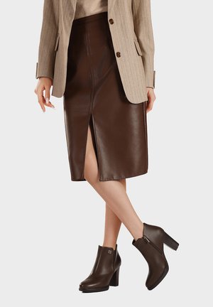 Donna che indossa una gonna in pelle marrone con spacco frontale, blazer color cammello e stivaletti marroni scuri con tacco, in piedi con le gambe incrociate.