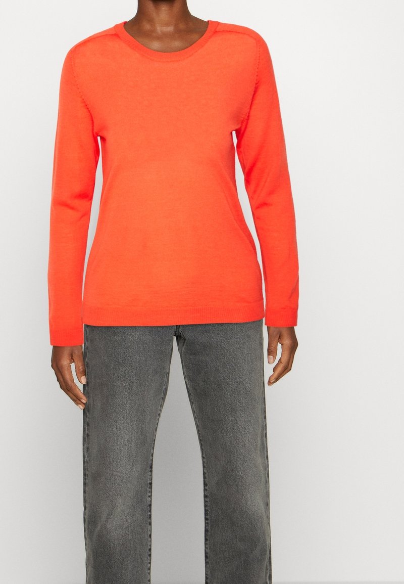 Pull en tricot orange avec un col rond, des manches longues et des détails côtelés à l'ourlet et aux poignets, associé à un jean droit gris.