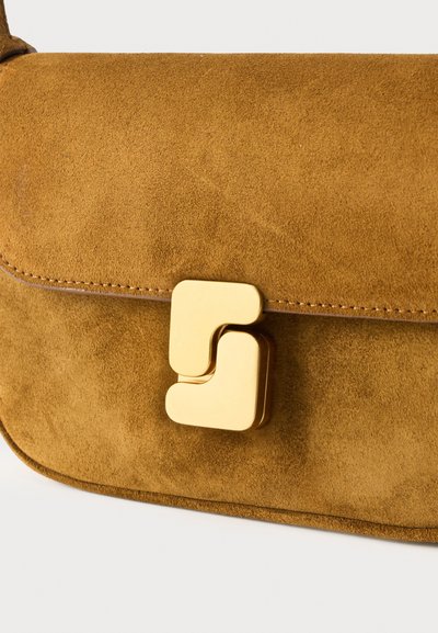 Μπεζ τσαντάκι ώμου από σουέντ με στρογγυλεμένες άκρες και χαρακτηριστικό χρυσό γεωμετρικό κούμπωμα στη μπροστινή πτέρυγα.