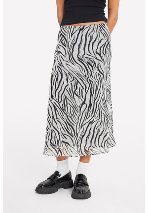 Osoba ubrana w czarny top, spódnicę o długości do kostek w nadruk zebry, białe skarpetki oraz czarne platformowe loafersy stojąca na białym tle.