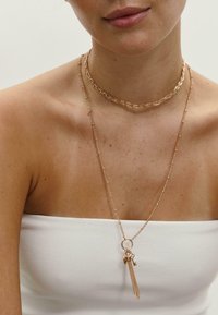 Guldtonede lagdelte halskæder med en snoet choker og en vedhæng med frynser og charme, mod bar hud med en hvid top.