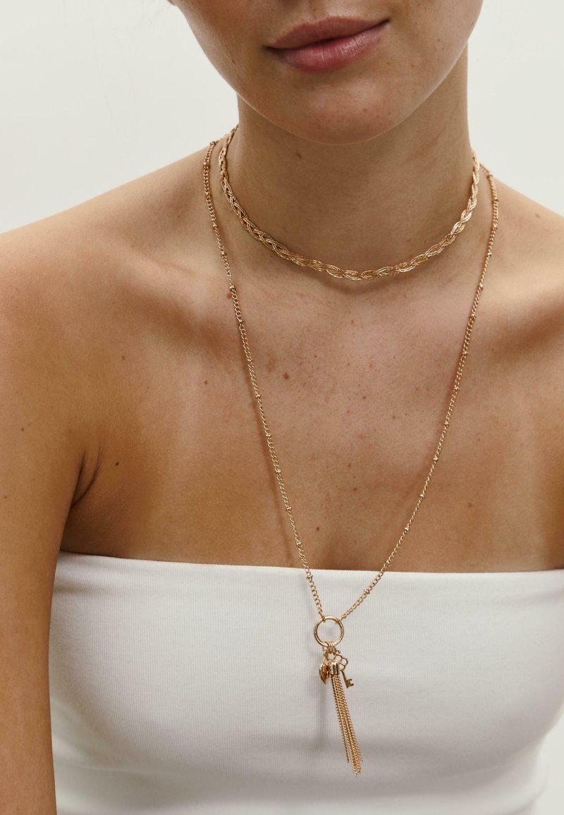 Guldtonede lagdelte halskæder med en snoet choker og en vedhæng med frynser og charme, mod bar hud med en hvid top.