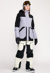 Snowboardjakke i lys lilla med sorte detaljer, der har en lynlåset front, store lommer og hætte; parret med sorte og cremefarvede bukser.