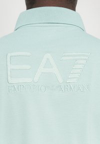 Gaiši zila polo krekla ar izšūtu tekstu "EA7 Emporio Armani" uz muguras. Mīksta tekstūra ar klasisku apkakles dizainu.