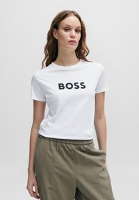 White cotton t-shirt featuring "BOSS" in bold black letters. Paired with loose, olive green pants, creating a casual ensemble.