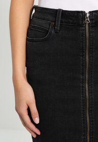 Falda de mezclilla negra con cremallera frontal, dos bolsillos laterales y una silueta ajustada. Textura suave con un brillo sutil.