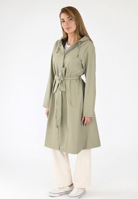 Trench-coat vert clair avec capuche, taille ceinturée, manches longues et poches latérales. Porté par-dessus un pantalon couleur crème et des baskets blanches.