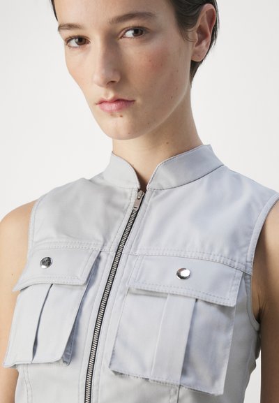 Sleeveless, light gray utility dress with a high collar, front zipper, and two chest pockets featuring snap buttons. Smooth fabric texture.