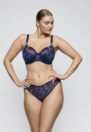 Vrouw die een bijpassende marineblauwe kanten bh en slip draagt met bloemmotieven, staat met haar handen in haar zij, kijkt opzij tegen een effen achtergrond.