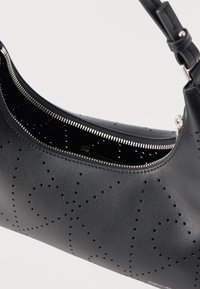 Μαύρη δερμάτινη τσάντα ώμου με διάτρητο σχέδιο, ασημένιο φερμουάρ ελαφρώς ανοιχτό και ρυθμιζόμενο λουρί, μπροστά σε απλό φόντο.