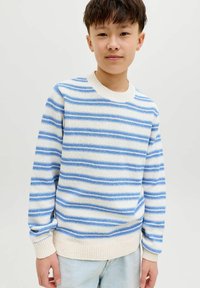 Garçon portant un pull en tricot rayé blanc et bleu et un jean bleu clair, se tenant devant un fond uni.