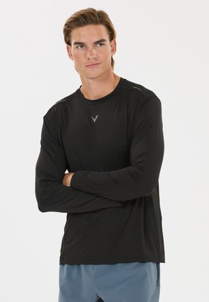Junger Mann mit hellbraunen Haaren, der ein schwarzes Langarmshirt und blaue Hosen trägt, die Arme verschränkt, schaut leicht nach rechts vor weißem Hintergrund.