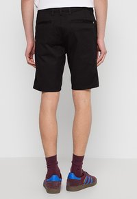 Homme portant un short noir jusqu'aux genoux, des chaussettes bordeaux et des baskets bordeaux avec des bandes bleues, debout devant un fond clair uni.