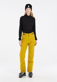 Zwarte zip-up top, levendige gele skibroek met hoge taille en zwarte knoopaccenten, gecombineerd met zwarte skischoenen en een zwarte beanie.