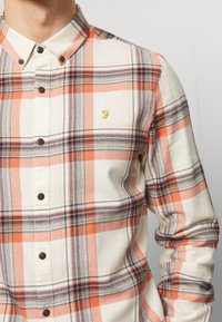 Geruit button-up overhemd in crèmekleurig, oranje en blauw; heeft een zachte textuur, kraag en een klein gouden logo op de borst. Bruine knopen.