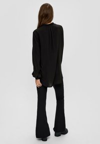 Zwarte blouse met lange mouwen en subtiele plooien, gecombineerd met zwarte flared broek en stevige zwarte laarzen, alles tegen een neutrale achtergrond.