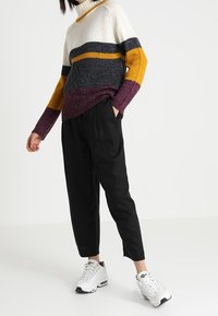 Strickpullover in Color-Blocking mit weißen, senfgelben, grauen und kastanienbraunen Streifen, kombiniert mit schwarzen, zulaufenden Hosen und weißen Sneakers.