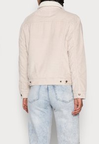 Beige kordjacka med ett shearling-krage, knäppte ärmslut och vertikal ribbad struktur; bärs över ljusblå denimjeans.