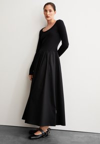 Schwarzes langärmeliges Kleid mit eng anliegendem Oberteil und ausgestellt geschnittenem, knöchellangen Rock. Kombiniert mit schwarzen Schuhen mit genoppten Riemen. Glatte Textur.