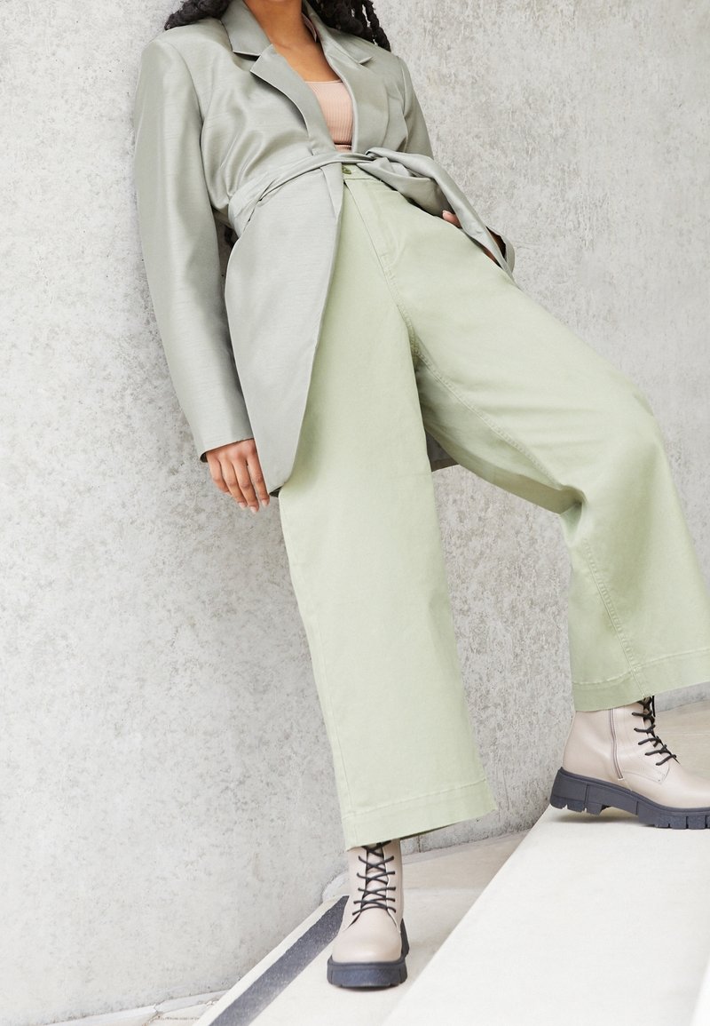 Femme portant un pantalon large vert clair, un blazer assorti ceinturé, des bottines beiges à lacets, debout sur des marches blanches contre un mur gris.