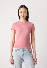Světle růžové bavlněné tričko s kulatým výstřihem, krátkými rukávy a bílým logem "TOMMY JEANS" na hrudi. Nosené s světle modrými roztrhanými džíny.