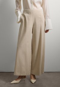 Femme portant un pantalon beige à jambes larges, une blouse blanche à manches longues avec de larges poignets, et des escarpins à talons kitten beiges, se tenant devant un mur uni.