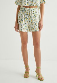 Shorts blancs avec un motif de citrons et de feuilles vertes, dotés d'une taille élastique et d'un cordon de serrage. Associés à des talons verts à brides.