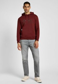 Czerwona bluza z kapturem z kieszenią kangurką, z nadrukowanym czarnym logo, w połączeniu z jasnoszarymi dżinsami i kremowymi trampkami. Gładka tekstura materiału.
